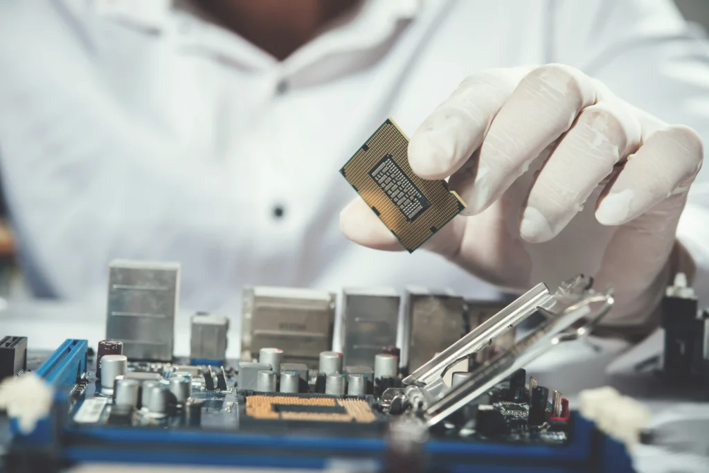 A technician wearing protective gloves holds a computer processor above a motherboard with the CPU socket and cooling components visible, representing the process of applying thermal paste to a CPU.