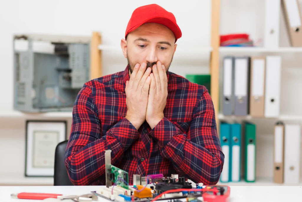 Concerned computer technician sitting at a workbench and looking at a desktop motherboard, with various computer components and tools visible in front of him.