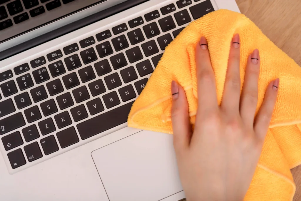 Close-up of a person cleaning a laptop keyboard and surface with a yellow microfiber cloth.