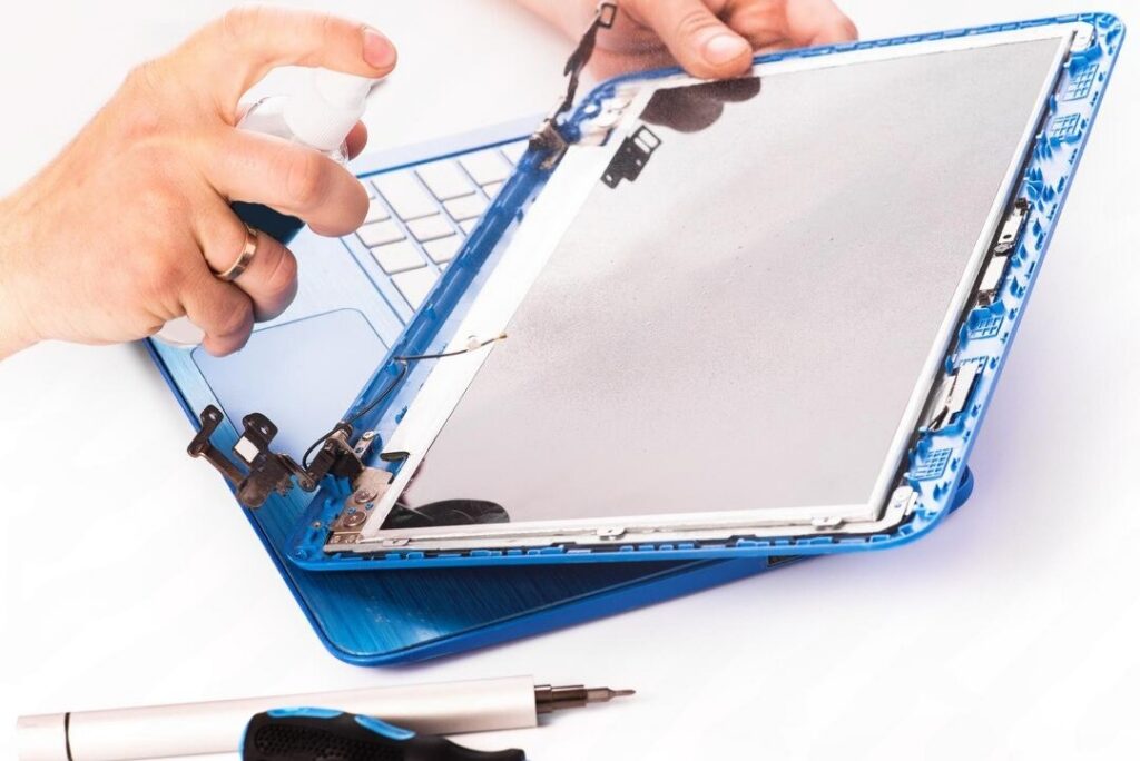 A technician disassembling a laptop and cleaning or repairing its exposed display panel with tools beside it.