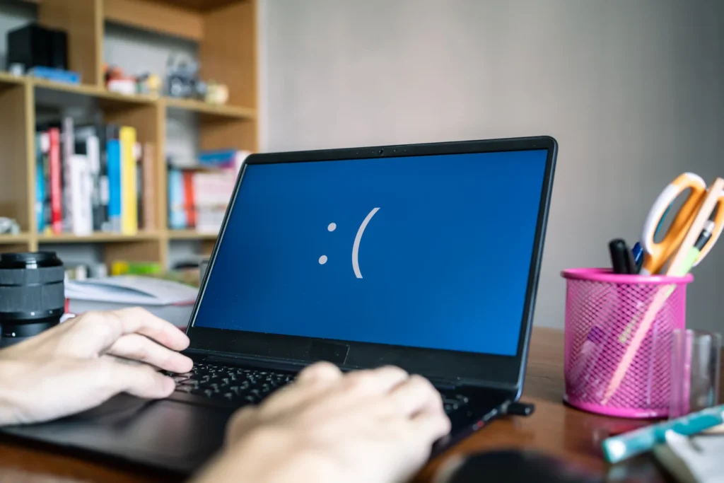 Hands are typing on a laptop showing a sad face emoticon on a blue screen, indicating an error. A cluttered desk and bookshelf are in the background.