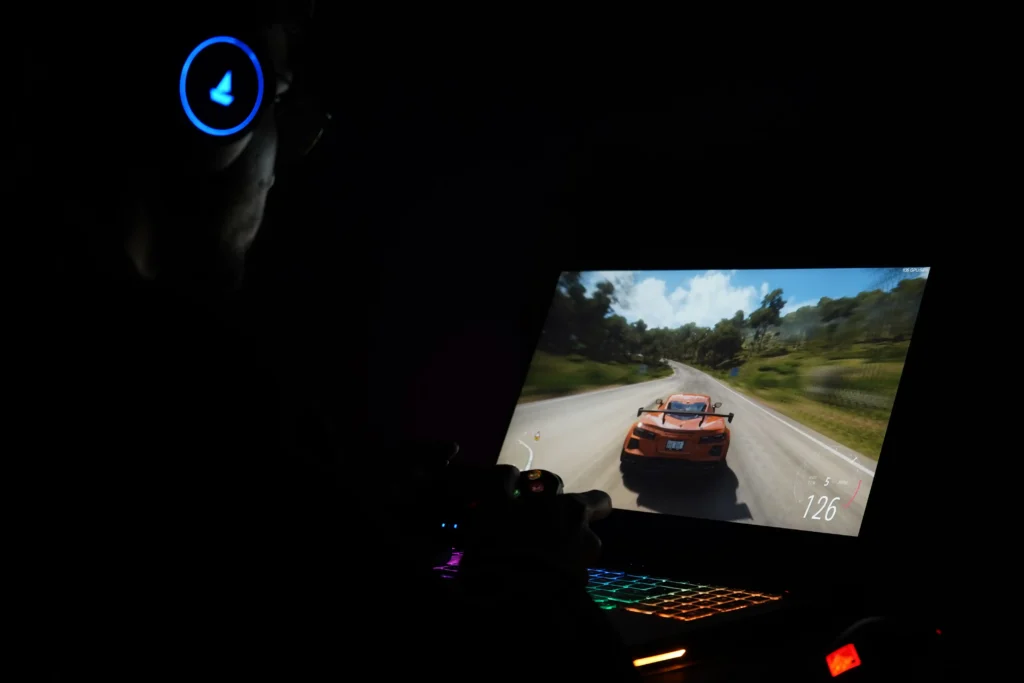 A person using a gaming laptop in a dark room while playing a racing game, with the illuminated keyboard and screen visible.
