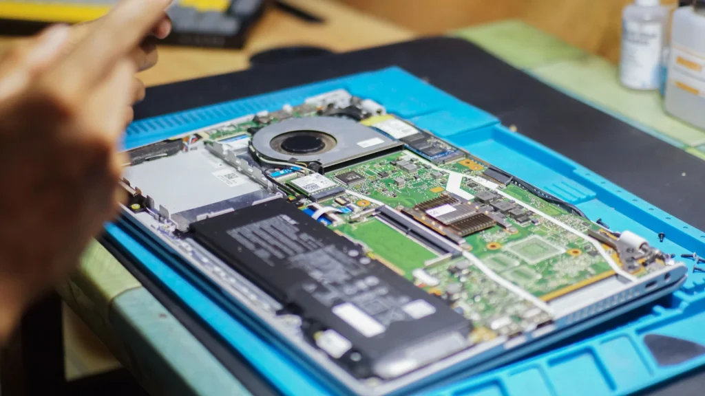 A technician inspecting the internal components of an opened laptop, with the battery, cooling fan, motherboard, and other hardware exposed for repair.