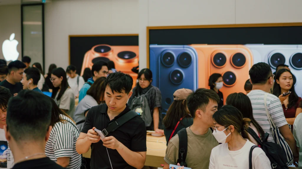 A spacious, modern store with rows of laptops on a white table. People in Apple shirts and visitors focus on the products, creating an excited buzz.