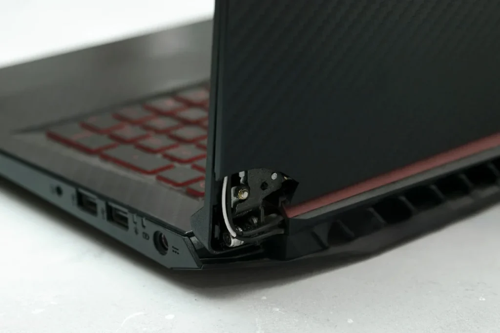 Close-up of a laptop with a damaged hinge exposed at the corner, showing the broken hinge mechanism, internal components, screws, and surrounding casing separating near the screen and keyboard base. Select 69 more words to run Humanizer.