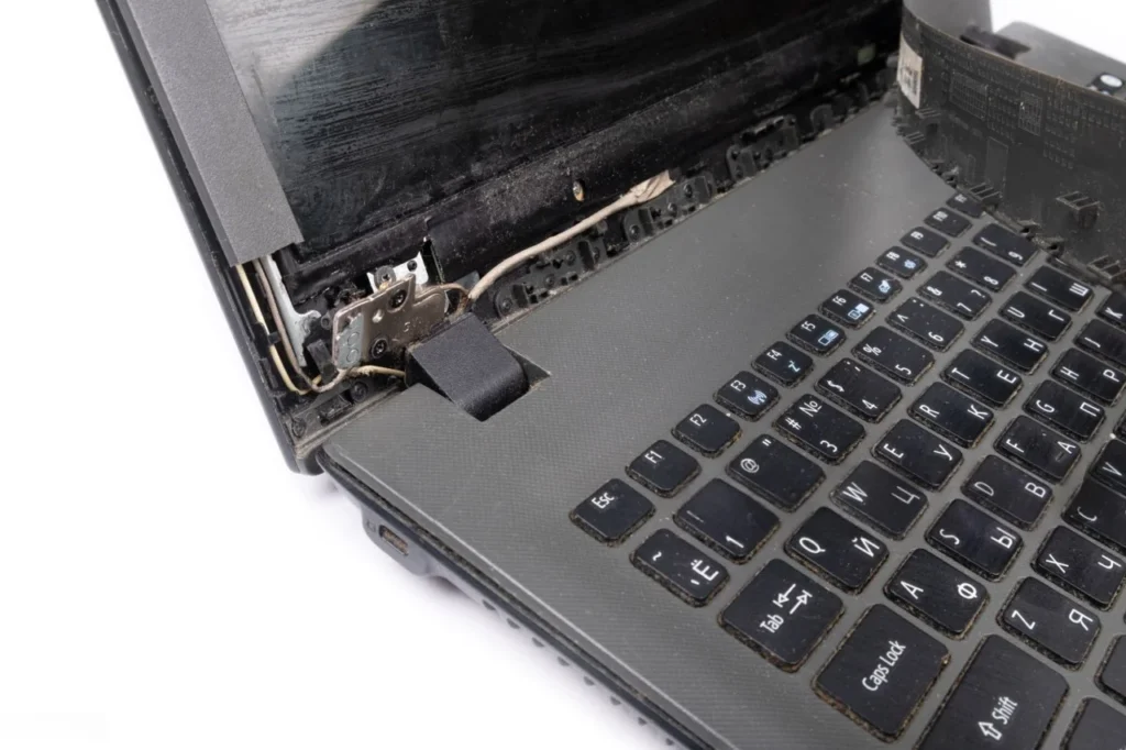 Close-up of a laptop with a severely broken hinge, showing the exposed metal hinge mechanism, internal cable, damaged casing, and separation between the screen and keyboard base.