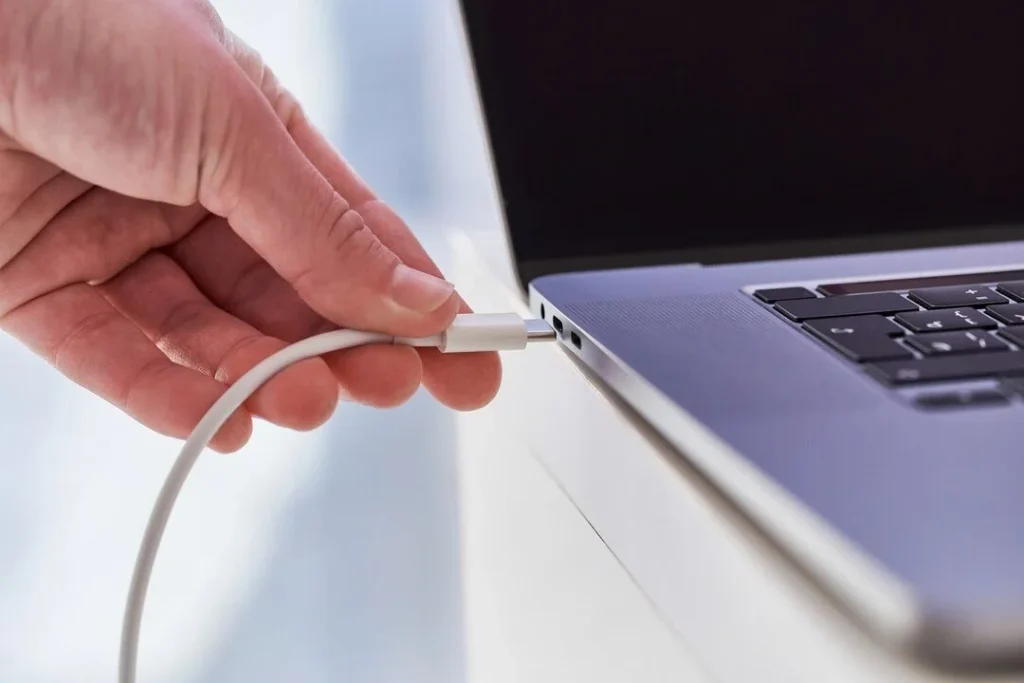 Hand connecting a charging cable to a laptop power port.