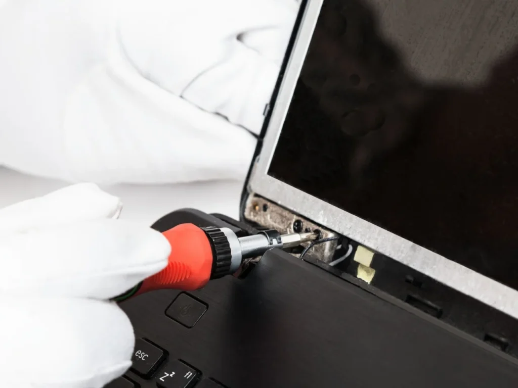 Close-up of a technician using a screwdriver to repair the hinge area of a partially disassembled laptop, with the display frame opened to expose the hinge mechanism and internal components. Select 69 more words to run Humanizer.
