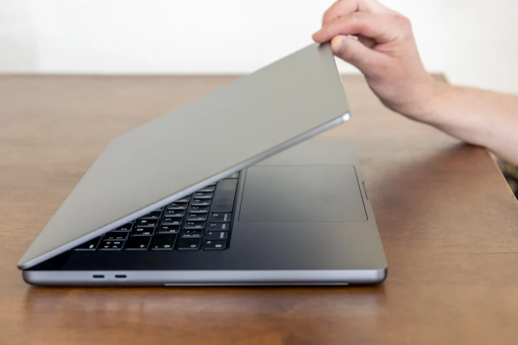 Side view of a person opening a slim gray laptop by lifting the display from the keyboard base, showing the laptop hinge area as the screen is adjusted on a wooden table.