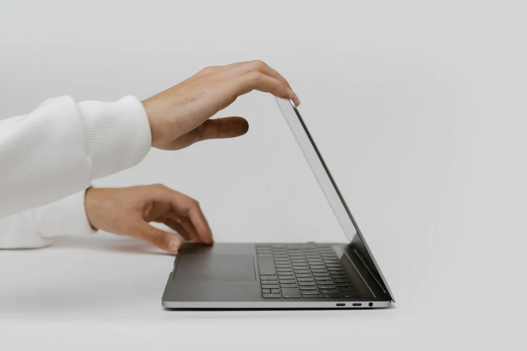 A person in a white sweatshirt gently opens a slim gray laptop, with one hand holding the display and the other steadying the base, showing the laptop hinge as the screen is adjusted.